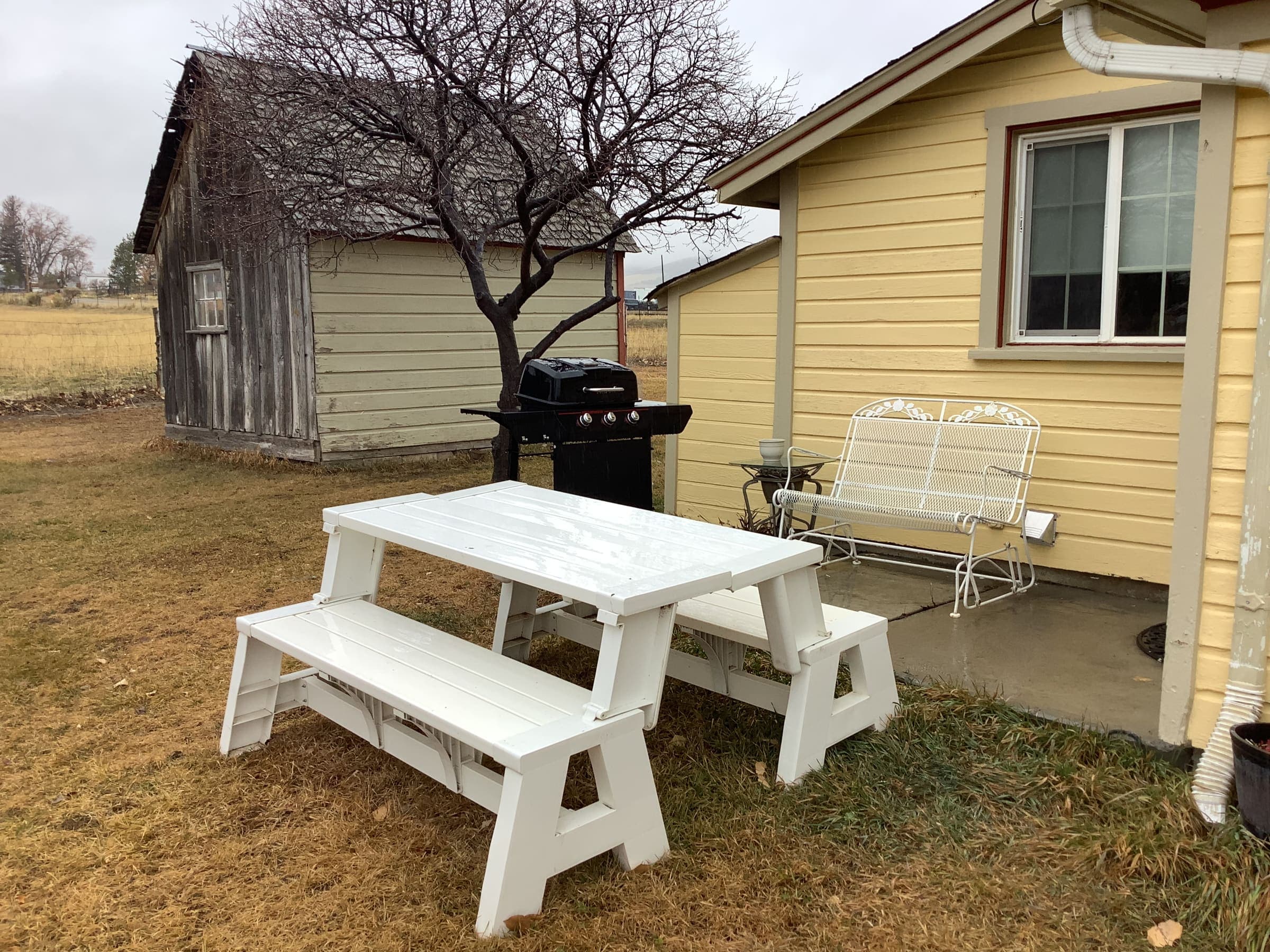 White picnic table and porch swing on the back lawn of the farmhouse, with the grill and old shed nearby